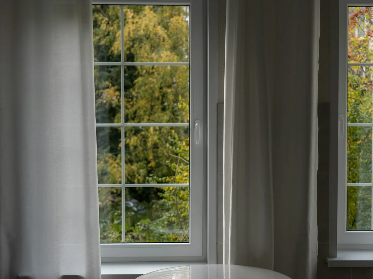 Tall residential windows looking onto trees, with light curtains drawn back